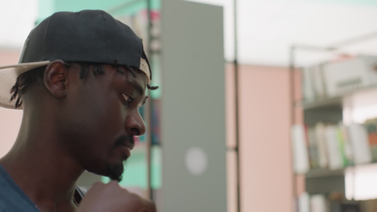 Close up of black man in casual cap holding stack of books in library, glancing thoughtfully at shelf while touching beard, suggesting study