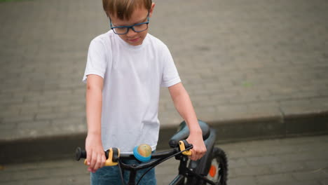 a young boy in glasses looks down as he holds the brakes of his bicycle while rubbing his head against his shoulder