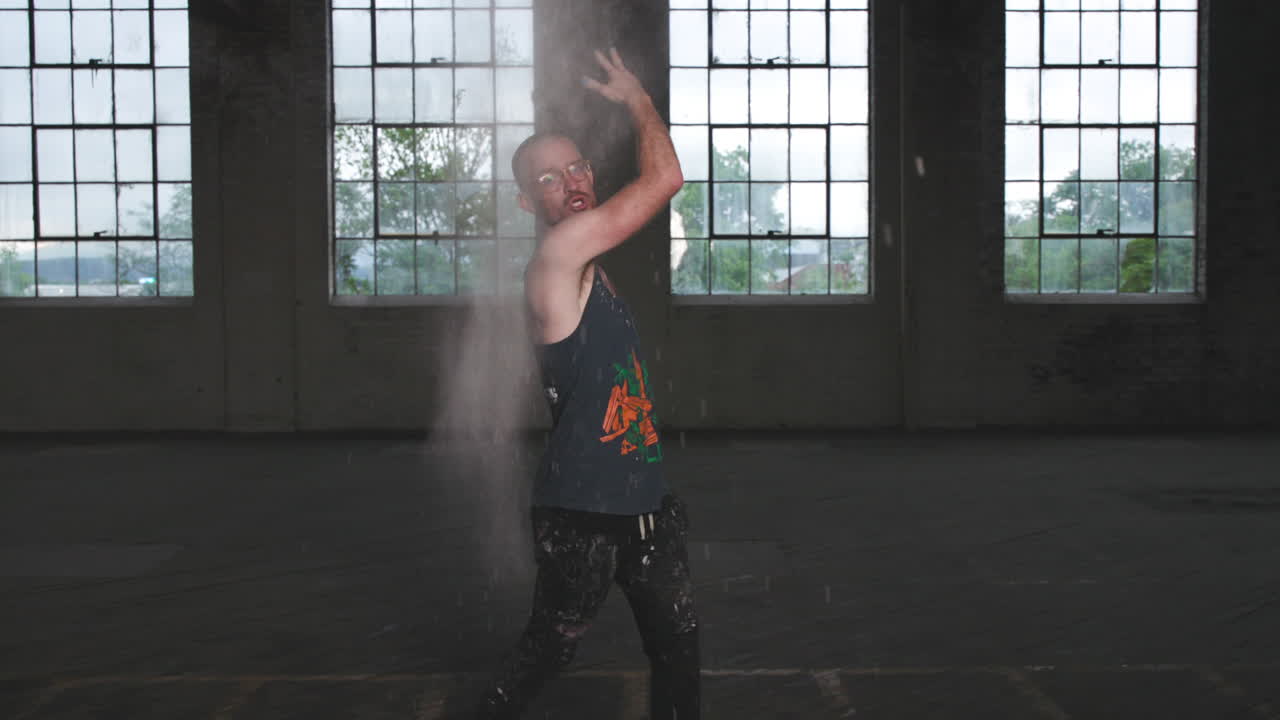 Man in an industrial warehouse in a tank top throwing flour in the air and being expressive with his face.