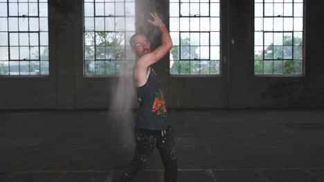man in an industrial warehouse in a tank top throwing flour in the air and being expressive with his face.