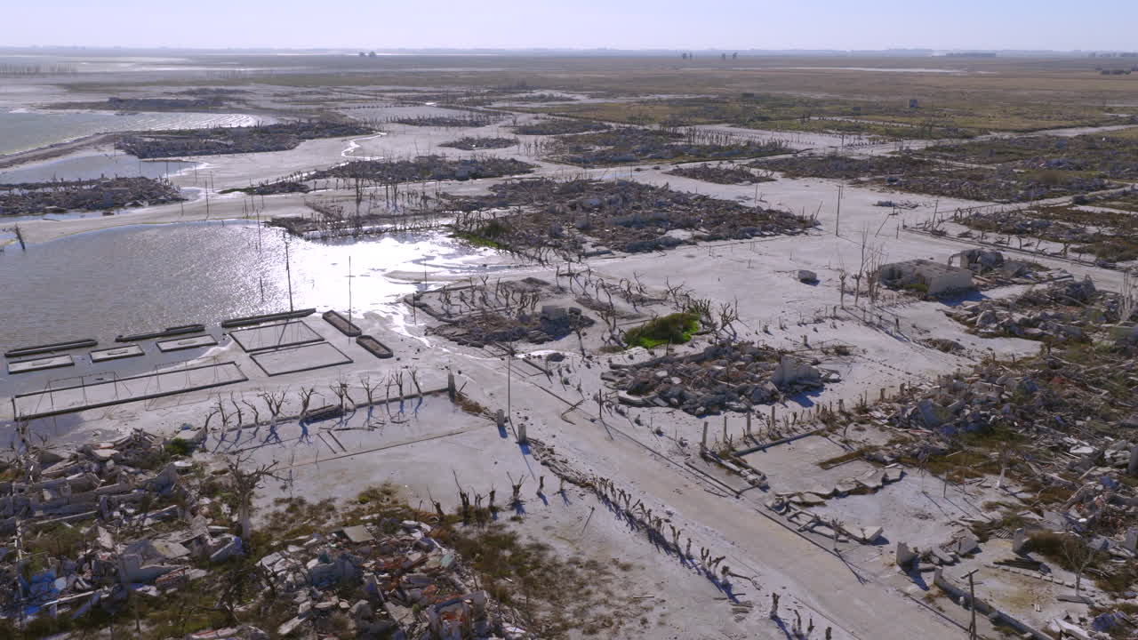 Aerial slow pan reveals Villa Epecuén geometric street grid, skeletal utility poles, collapsed structures and gleaming salted ground spreading from lakeshore spa complexes toward pampas, Argentina
