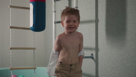 smiling toddler boy takes off torn shirt standing in gym