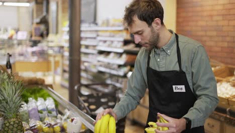 happy grocery store worker in apron caucasian bearded guy arranging bananas at modern organic supermarket grocery store