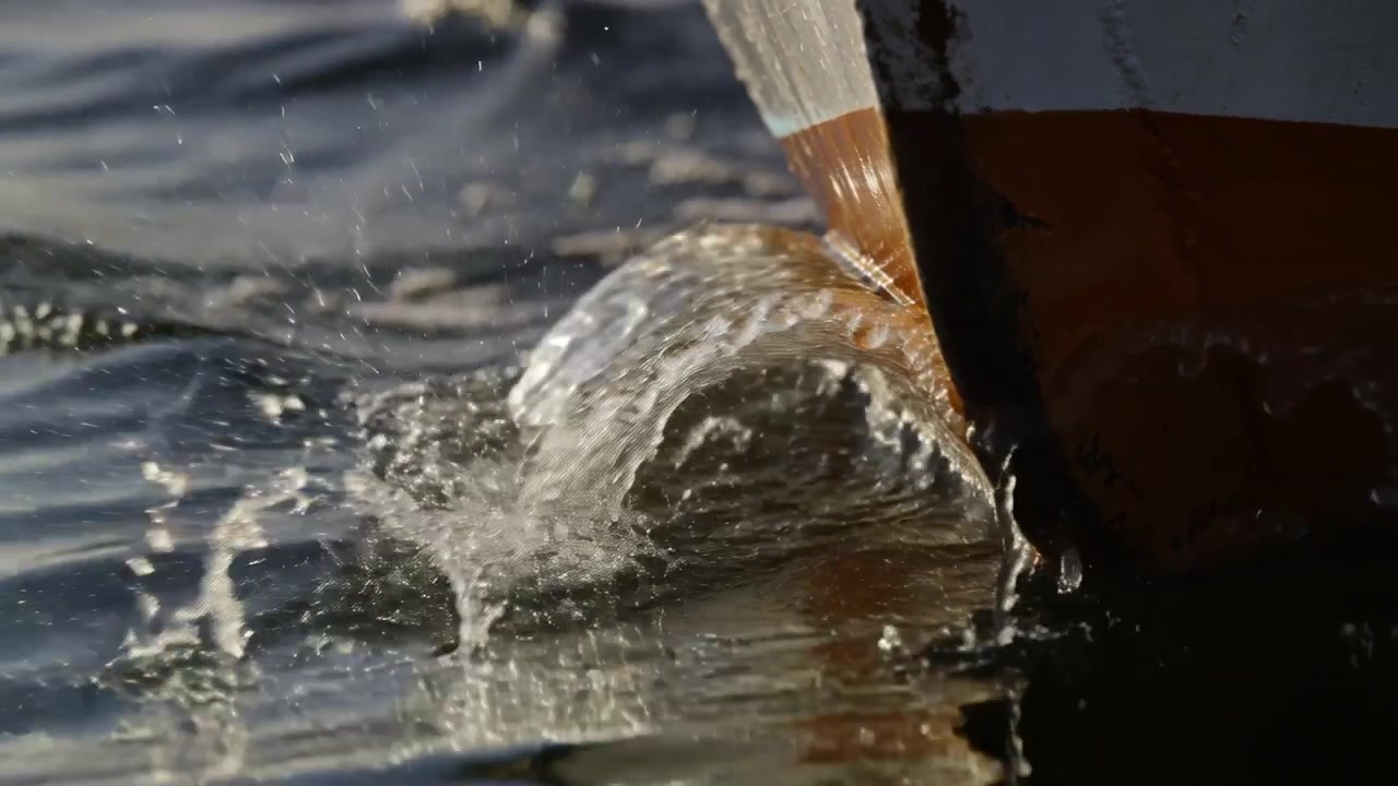 Prua verniciata che si immerge nell'acqua calma, formando un'onda di prua arricciata e riflessi del sole, con spazio per il testo.