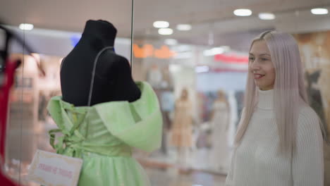 elegant lady walks to show glass admiring blue gown with gentle smile, other dresses and mannequins visible behind glass, blurred shoppers in background walking past bright boutique interior