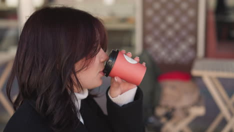 primer plano de una mujer con abrigo negro bebiendo café de una taza roja, sonrisa satisfecha mientras la coloca suavemente, con un fondo urbano borroso que mejora la sensación relajada y urbana