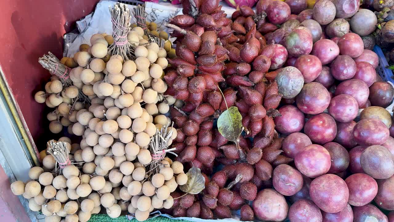 Camera pans over vibrant longan, snake fruit, passion fruit, and mangoes at a Thai market