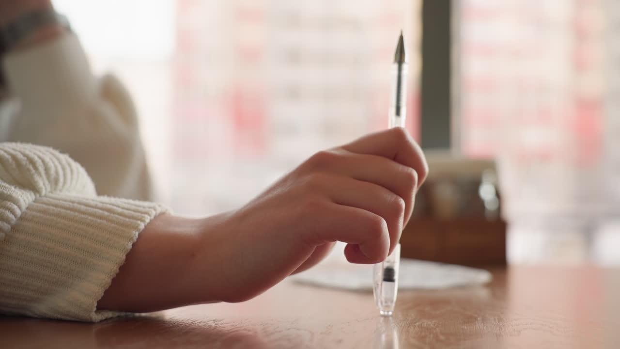 Close up hand holding pen and gently tapping it on wooden table beside napkin in soft natural indoor light suggesting thoughtful mood, waiting moment