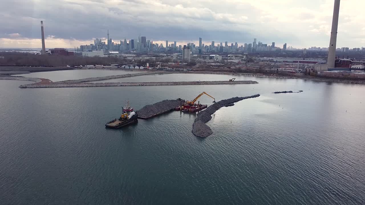 토론토 스카이라인을 갖춘 바지선에서 바위를 옮기는 굴착기에 의한 방파제 건설