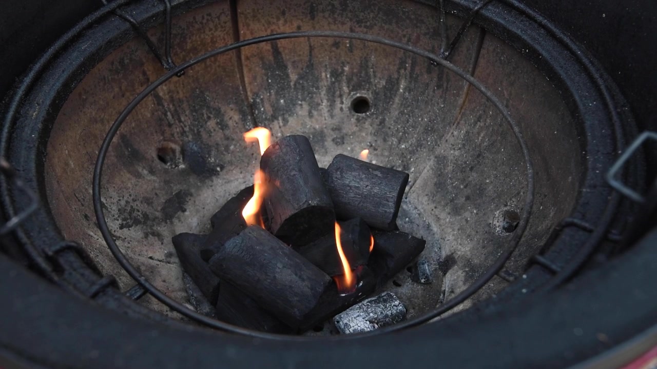 Detail hořícího kusového dřevěného uhlí v keramickém grilu typu kamado. Kovové kleště přesouvají uhlí, aby se rozšířily žhnoucí oranžové plameny.
