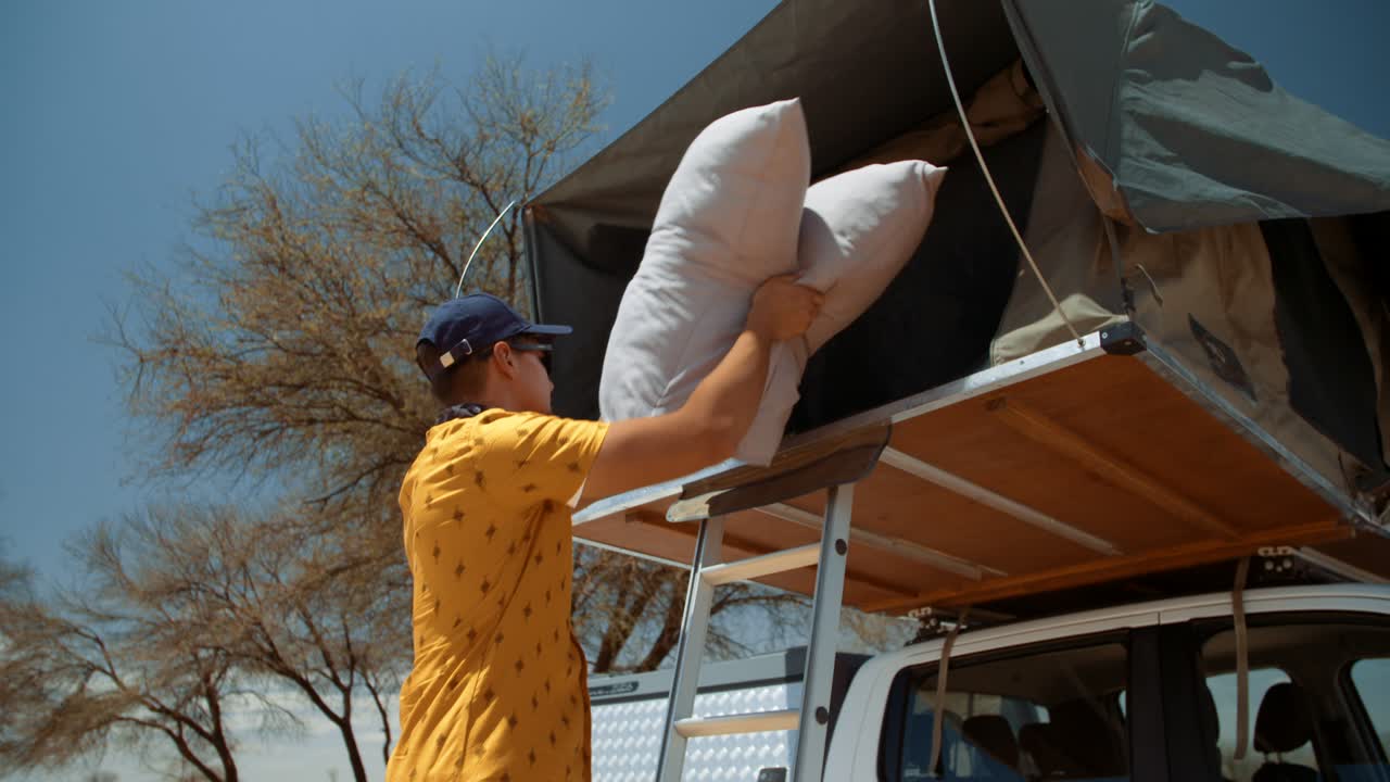 Hand held shot of a off-roading Caucasian male tourist in Africa as he walk towards a ladder of a rooftop tent while carrying pillows, he proceeds to put the pillows in his canvas tent and climb in.