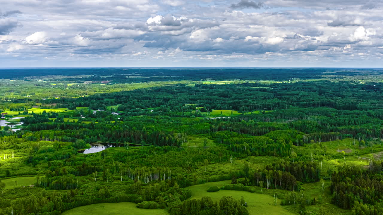 A dynamic aerial hyperlapse flies sideways at high speed over a vast and beautiful Latvian landscape, revealing a patchwork of green forests, fields, and pristine lakes