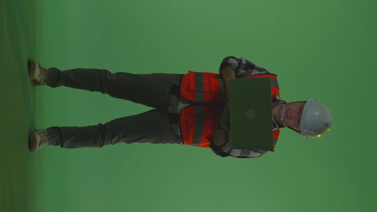 Full Body Of Asian Male Engineer With Safety Helmet Working On A Laptop And Looking Around While Standing In The Green Screen Background Studio, During Sunset Or Sunrise Time