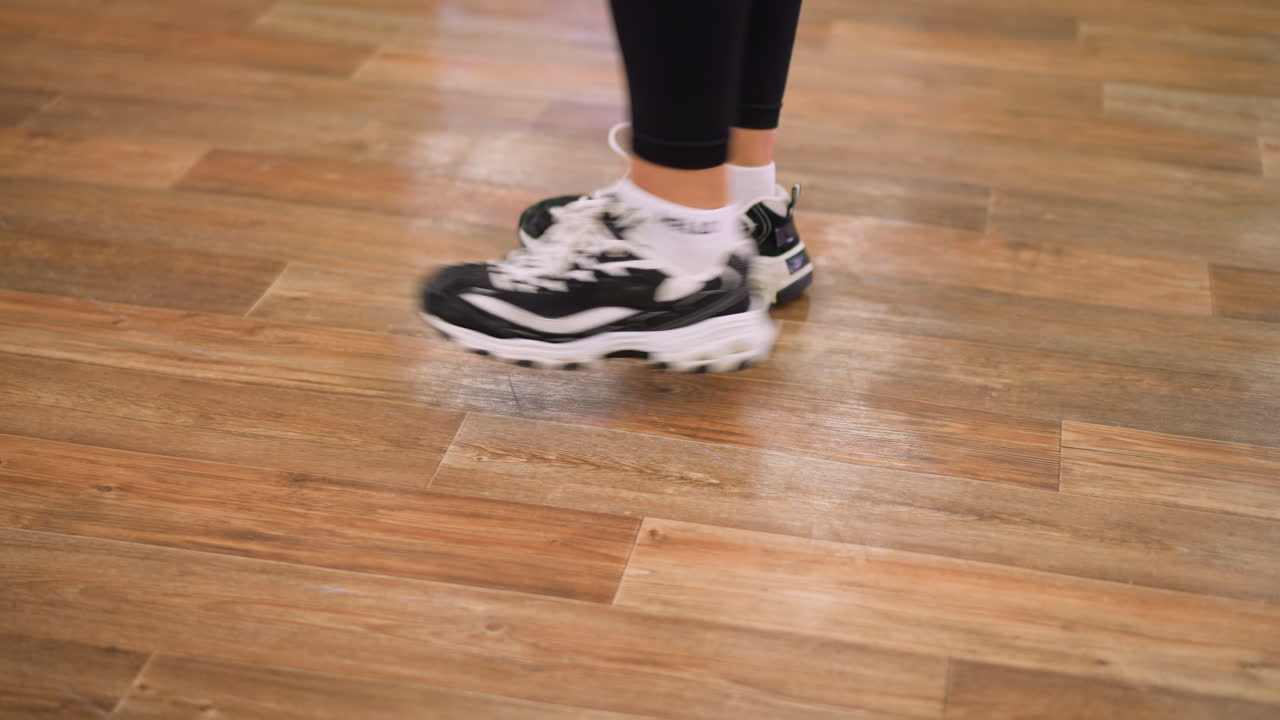 Side leg view in black leggings and canvas sneakers bouncing on polished wooden floor during active indoor workout, showing motion, rhythm, and strength through dynamic athletic expression