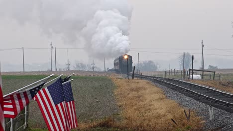 view of a steam passenger train approaching blowing smoke and steam with american flags waving in the wind on the fence on a cloudy day