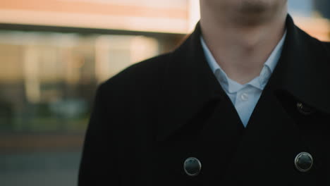 close-up of man standing with focused expression, wearing black coat and white shirt, soft glow of sunlight illuminating behind him, blurred background showcasing urban glass building