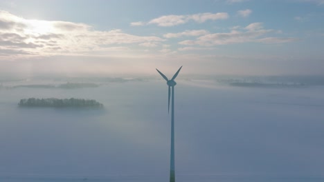 establishing aerial view of wind turbine generating renewable energy in the wind farm, snow filled countryside landscape with fog, sunny winter day, wide drone shot moving forward