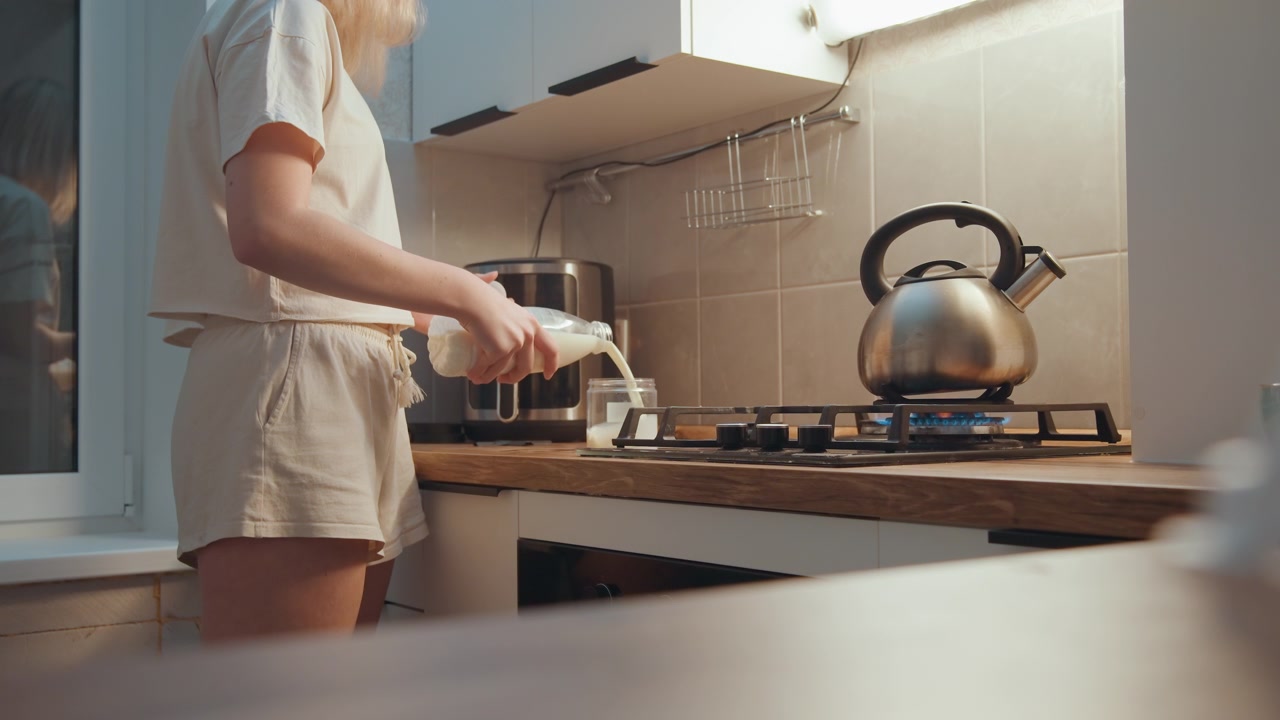 Cucina illuminata dal sole che versa il latte accanto al bollitore, preparazione della colazione mattutina con cartone del latte e macchina del caffè sul bancone, abbigliamento casual da casa, vapore dal bollitore, atmosfera accogliente e invitante e routine semplice