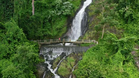 Joven-Asiática-Cruzando-Un-Puente-Con-Una-Poderosa-Cascada-Detrás-De-Ella,-Aérea