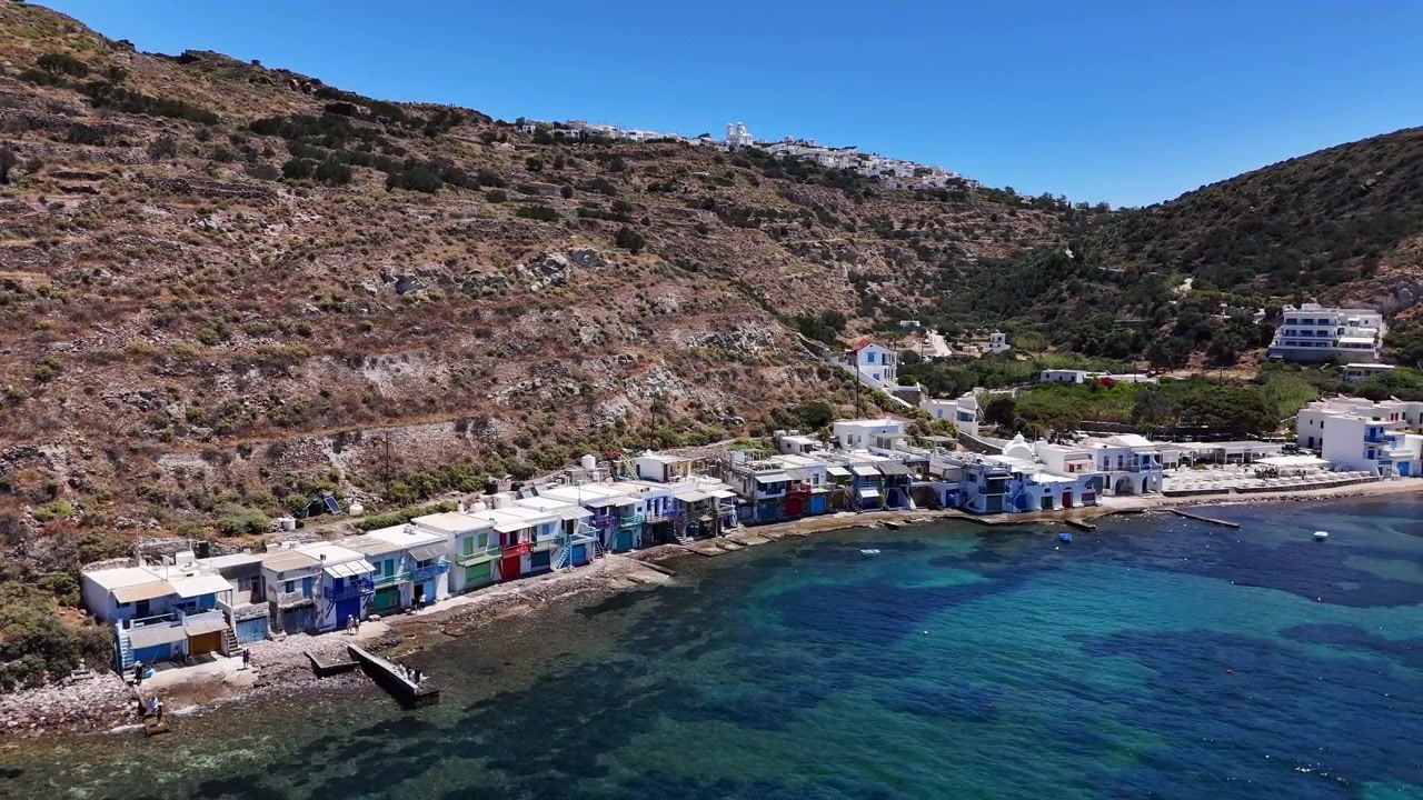 Colorate rimesse per barche di Klima passano sotto una vista aerea lungo la costa di Milos, con moli in legno, acqua bassa, case bianche e una valle verde sotto ripide colline terrazzate.