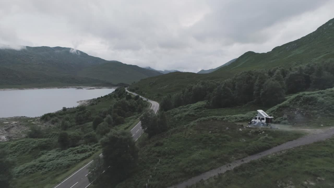 Wide push in drone shot of Loch Beag, traffic travelling along the coastal road and a campervan wild camping with BBQ smoke and mountains in the background in the Scottish Highlands