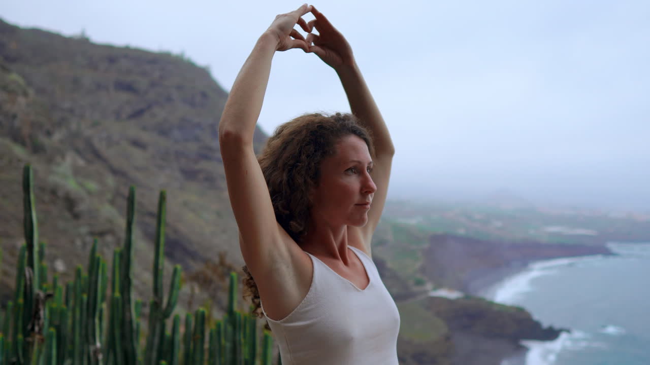 A woman meditates on a mountain's height, displaying the Maha Sakal hand gesture, while the ocean and green mountains provide a serene setting