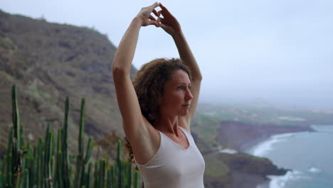 a woman meditates on a mountain's height, displaying the maha sakal hand gesture, while the ocean and green mountains provide a serene setting