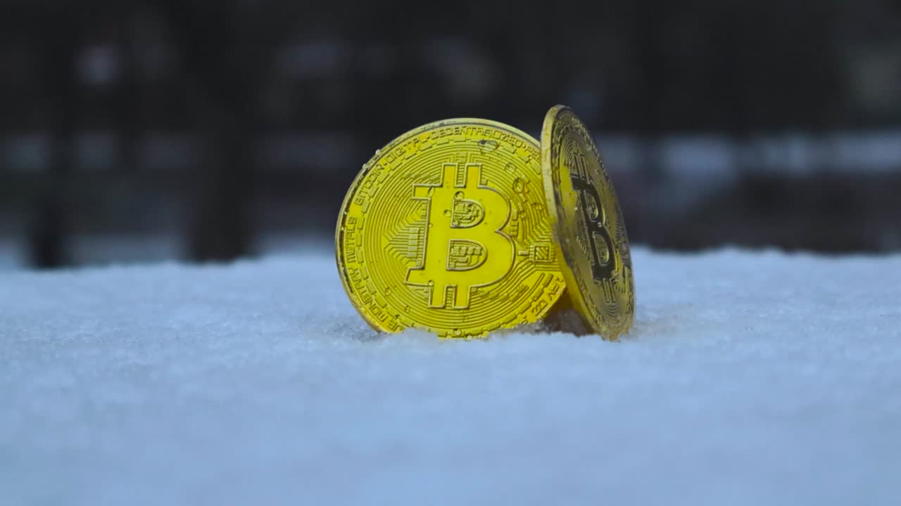 Two golden yellow reflective bitcoin cryptocurrency coins in display on a snow covered stand outdoors during day time while snow falls on them. Camera spins around coins while background is blurred.