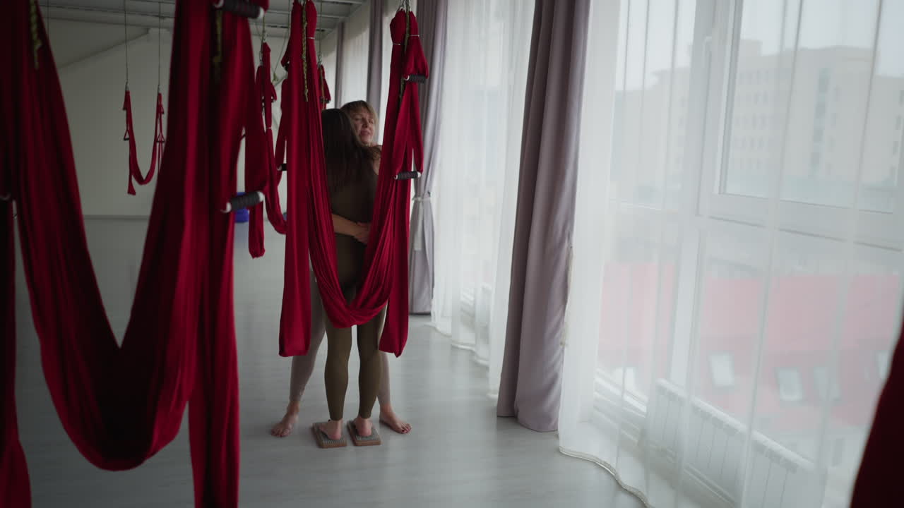 Yoga instructor embraces partner barefoot on nail board inside serene aerial silk studio under soft natural light beside mirrored wall and flowing curtains conveying healing support mindfulness