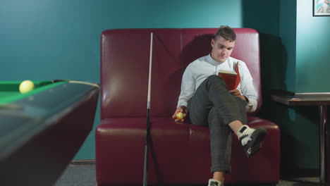 dude in white shirt and plaid pants sits on red sofa in billiard room, reading book while rolling orange billiard ball in hand. cue stick leans beside him. green pool table and dim lighting