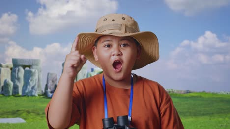 asian boy with a hat and binoculars thinking and looking around then raising his index finger while traveling in stonehenge. boy researcher, travel adventure concept, close up