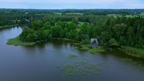 aerial shot with push in low angle shot over a lake with views of a countryside house nestled amongst a green forest in latvia