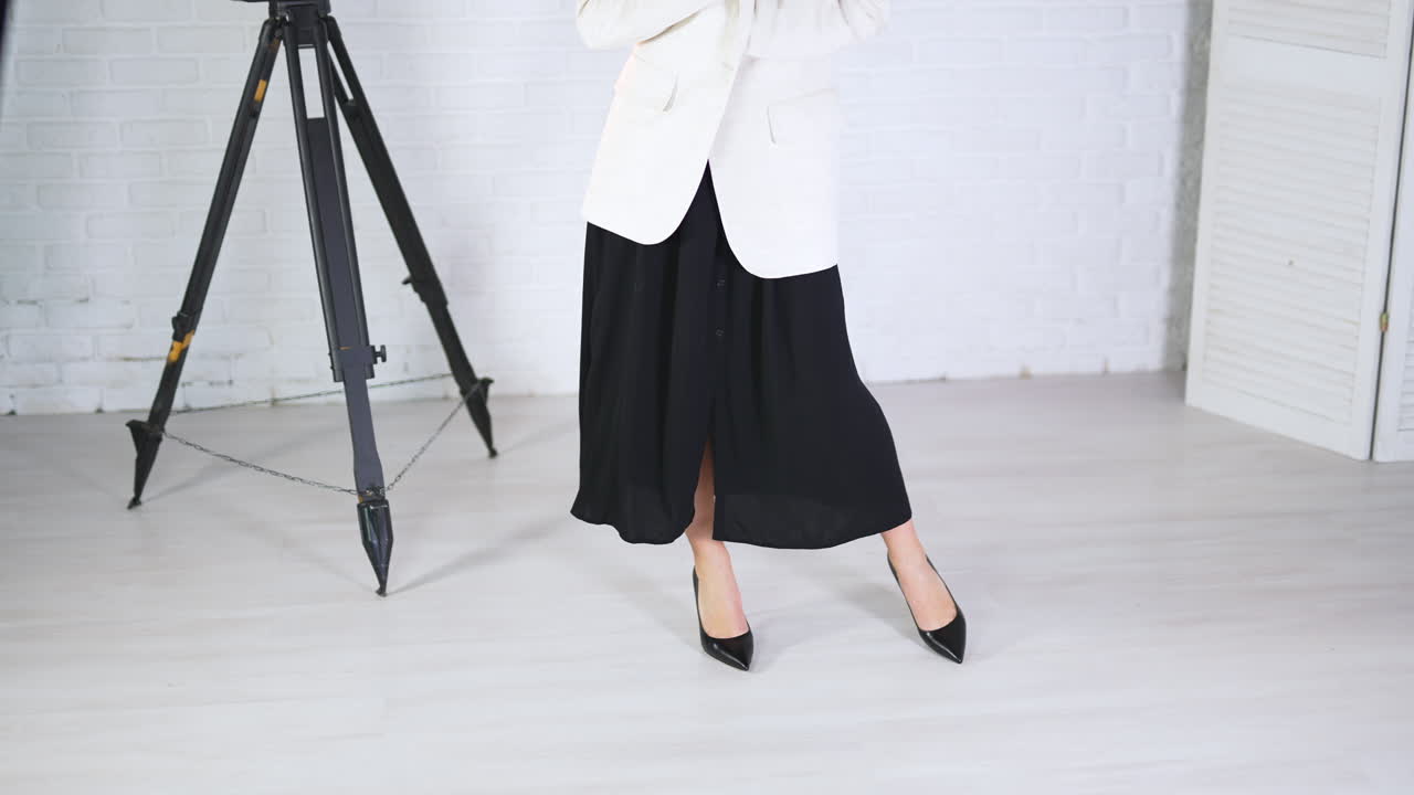Demonstrating black high-heeled shoes in studio. Woman in long dress and white jacket poses in classic shoes. White backdrop.