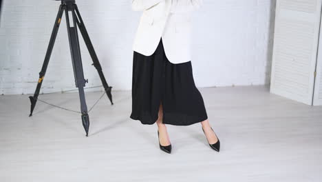 demonstrating black high-heeled shoes in studio. woman in long dress and white jacket poses in classic shoes. white backdrop.