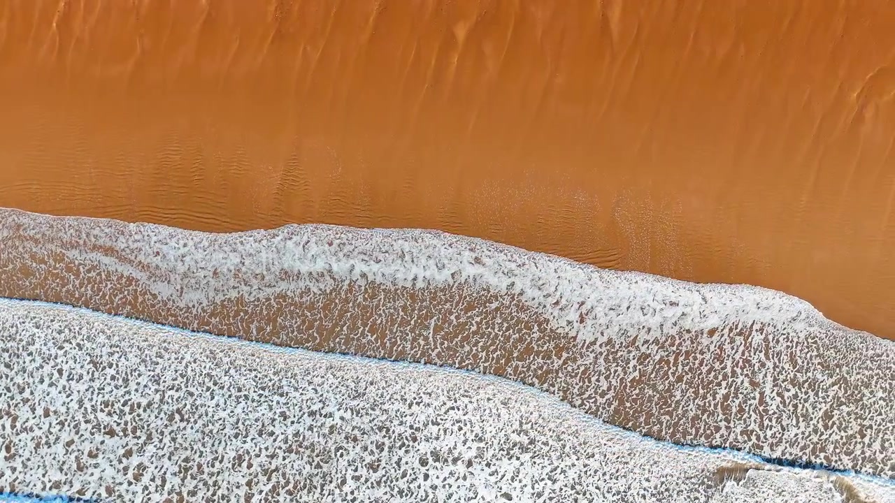 Abstrakt luftfoto af havbølger, der møder rustfarvet sandstrand. Et luftfoto ovenfra viser rustorange vådt sand afgrænset af et bånd af hvidt bølgeskum og en smal stribe blåt vand.