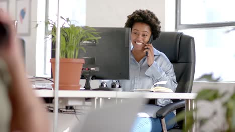 happy african american casual businesswoman talking on smartphone at desk in office, slow motion
