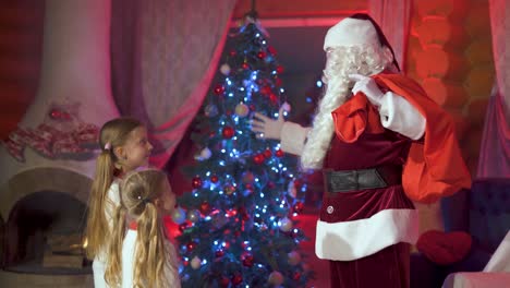 cheerful santa claus with a beard and a bag of gifts plays with little girls in a room with a christmas tree and glowing decorations