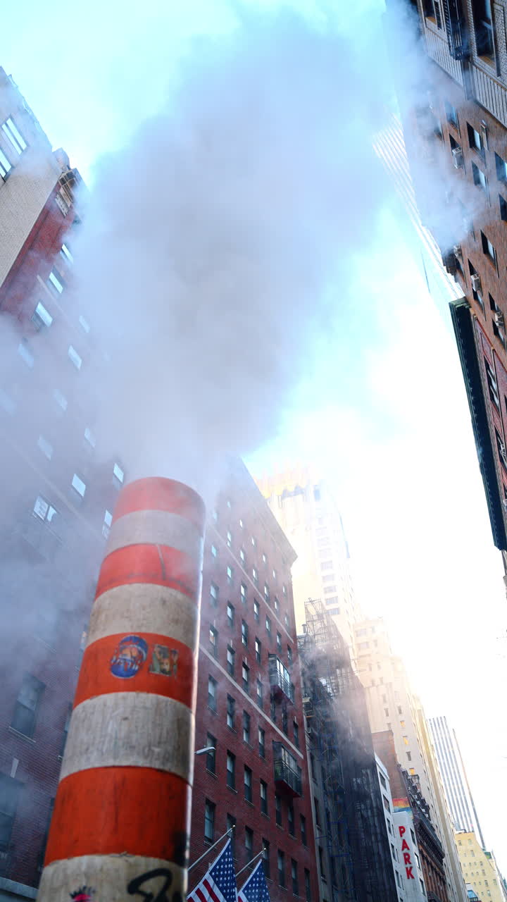 Red and white stripy tube releasing thick white steam outdoors. Low angle view. Multi-storied buildings of New York at backdrop. Vertical video