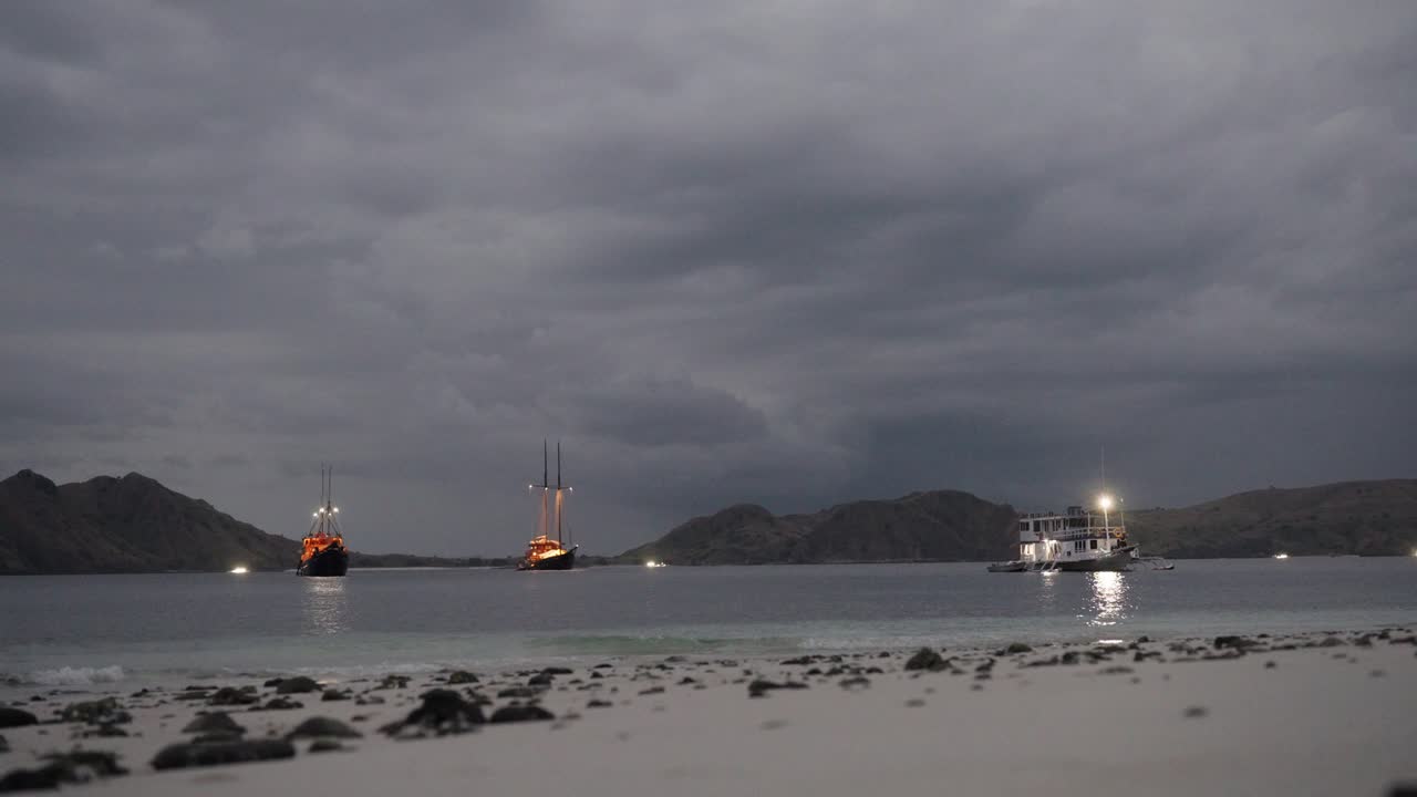 Fredeligt aftenbillede af oplyste skibe ankret nær stranden i Komodo Nationalpark med en mystisk skyet himmel og vulkanske bjerge i baggrunden