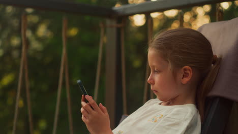 preschooler girl spends time playing mobile game on terrace. tranquil child uses smartphone reclining in comfortable armchair against wild summer forest