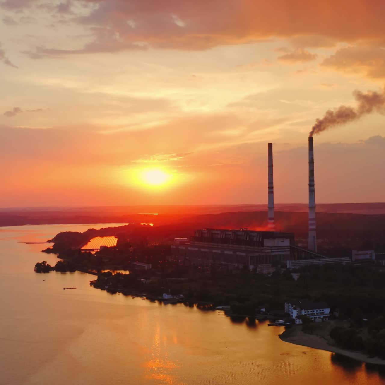 Industrial enterprise on orange sunset. Large plant with pipes near the river. Dirty smoke pours into the air from chimney. Environmental pollution. Aerial view.