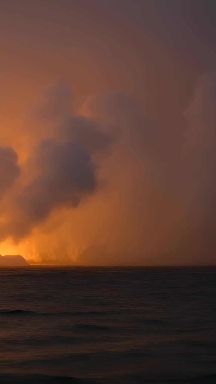 Vertical video: Bright shore glow sending smoke plume over ocean at dusk, lighting low clouds