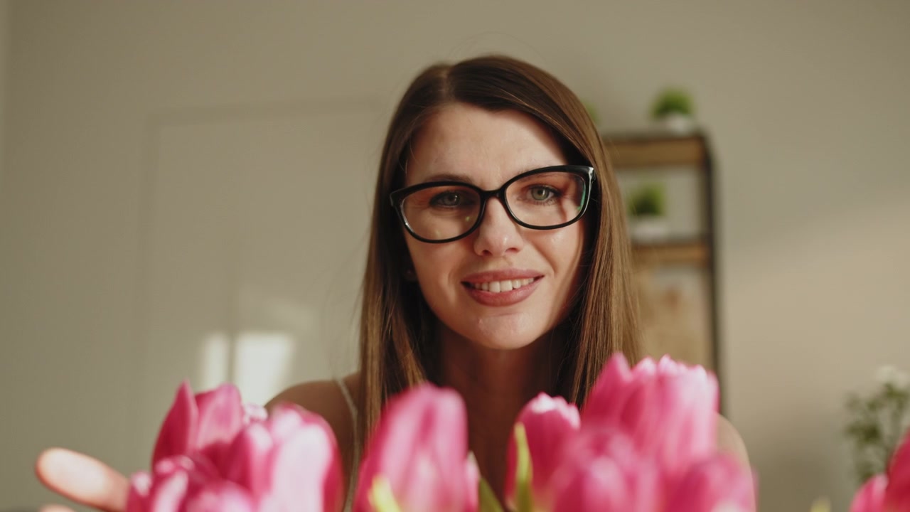Giovane donna con gli occhiali che tiene in mano un bouquet di tulipani rosa, sorridendo dolcemente in una stanza luminosa.