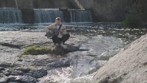 young man playing guitar sitting on the bank of a mountain river on a background of rocks. concept of freedom relaxation. place