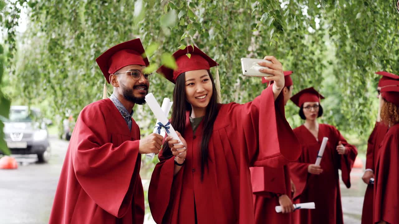 Fellow students are taking selfie with diplomas posing and smiling, girl is holding smartphone, people are wearing gowns and hats. Education and modern lifestyle concept.