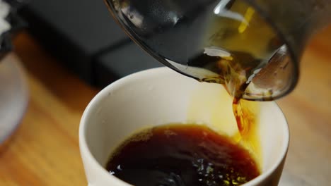 Closeup-shot-black-coffee-served-from-glass-jar-into-white-small-cup,-barista-shot-concept,-wooden-table-at-shop