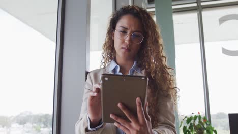 mujer de negocios biracial sentada en un escritorio y usando una tableta en interiores modernos