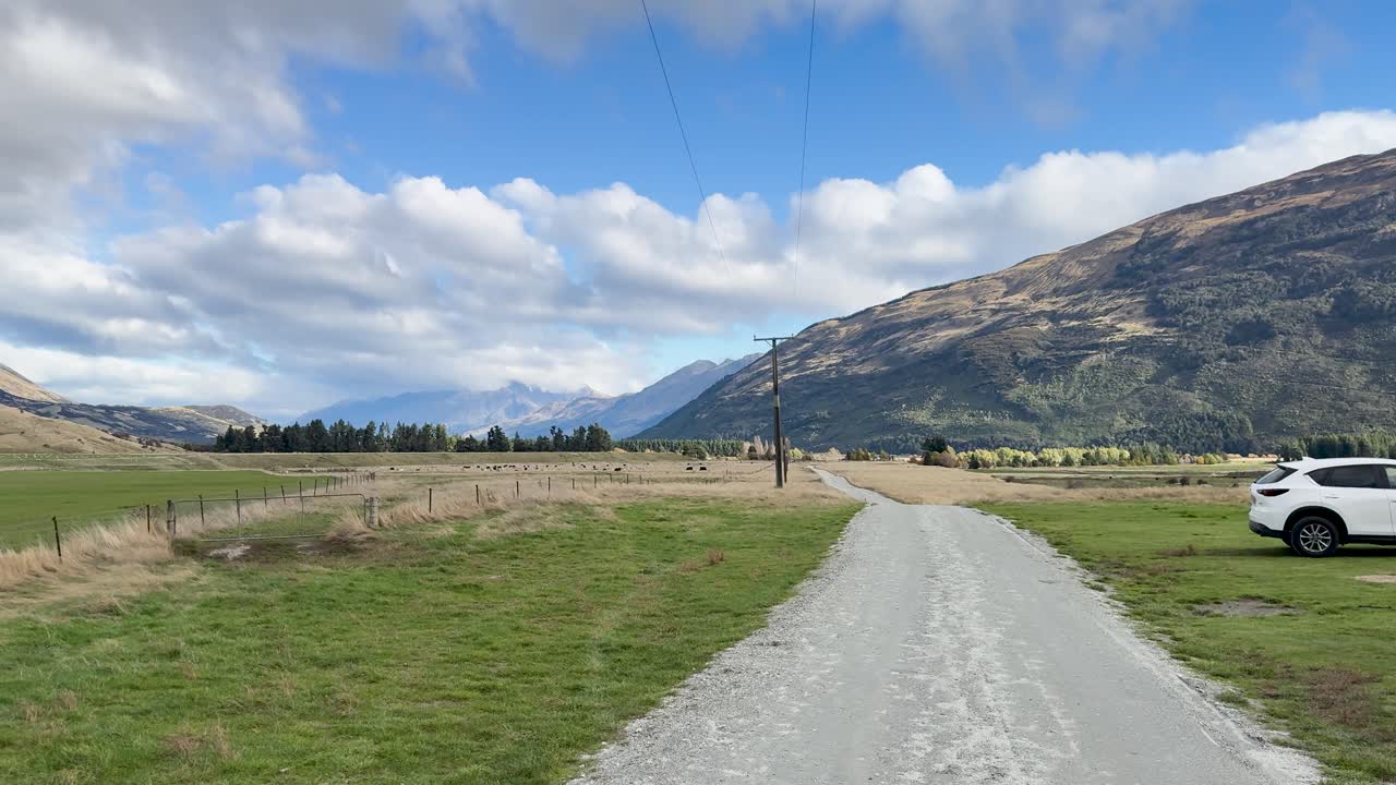 White SUVs drive on rural gravel road with mountains, grassy fields, and bright daylight