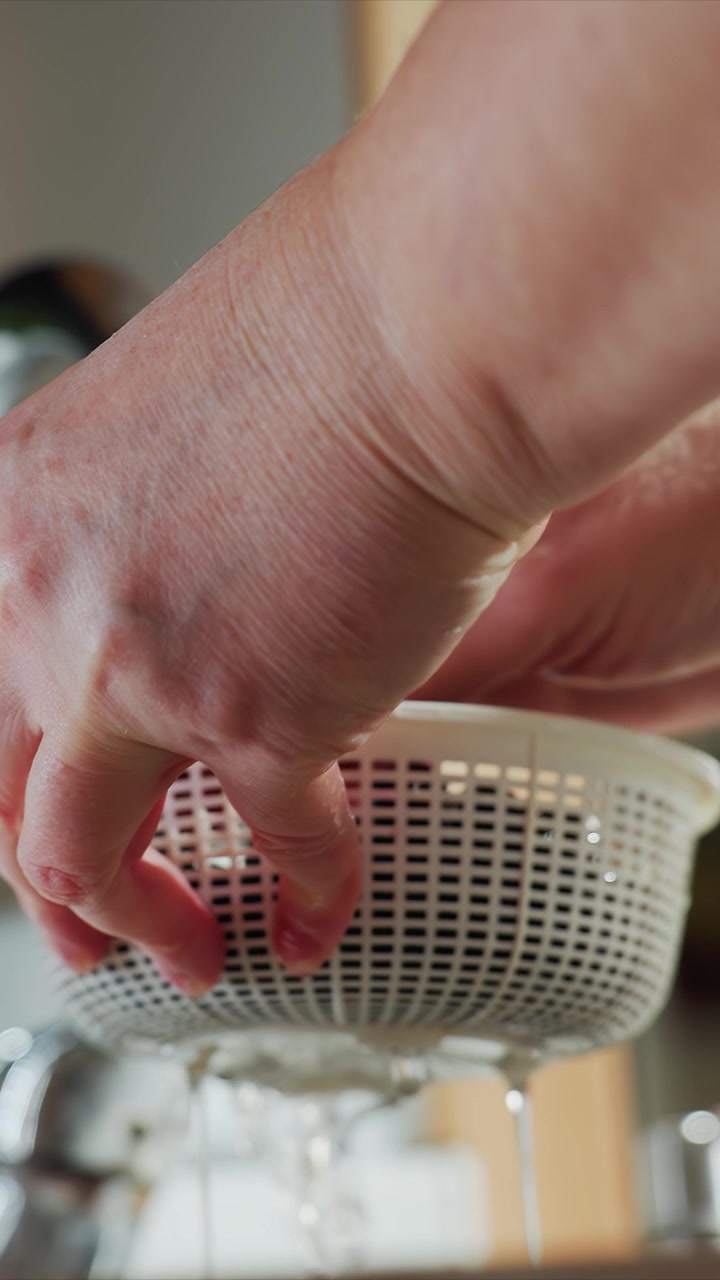 Close up of hands holding white plastic colander. Focus remains on motion and interaction between wet mesh strainer. Lighting is consistent indoors emphasizing foreground activity