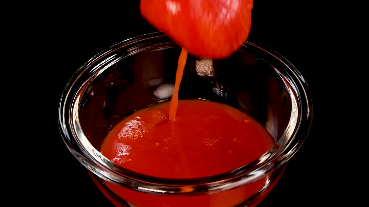 A whole tomato is dropped into a glass bowl filled with tomato sauce, creating a dramatic splash under bright studio lighting with a black background